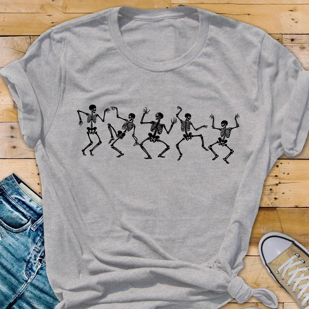 May include: A light gray t-shirt featuring a black graphic of five dancing skeletons. The skeletons are in various poses, with their arms and legs outstretched. The shirt is made of a soft material and has short sleeves.