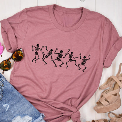 May include: Dusty rose-colored t-shirt featuring a black graphic of dancing skeletons. The skeletons are in various poses, with arms and legs outstretched. The shirt is made of a soft material and has a round neckline.