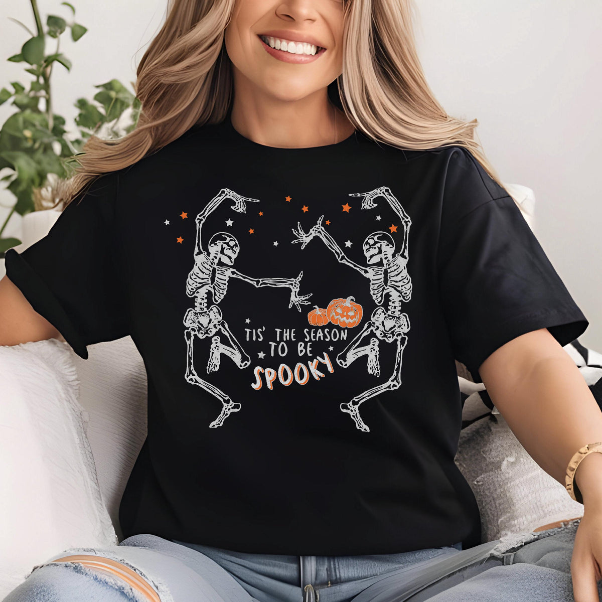 May include: Black t-shirt with a white skeleton graphic dancing with a pumpkin in the middle. The text "Tis' the season to be spooky" is printed below the skeletons. Orange stars are scattered above.