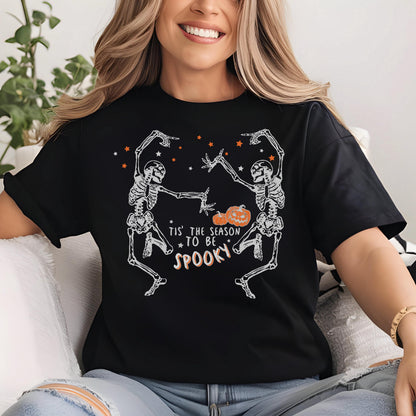 May include: Black t-shirt with a white skeleton graphic dancing with a pumpkin in the middle. The text "Tis' the season to be spooky" is printed below the skeletons. Orange stars are scattered above.