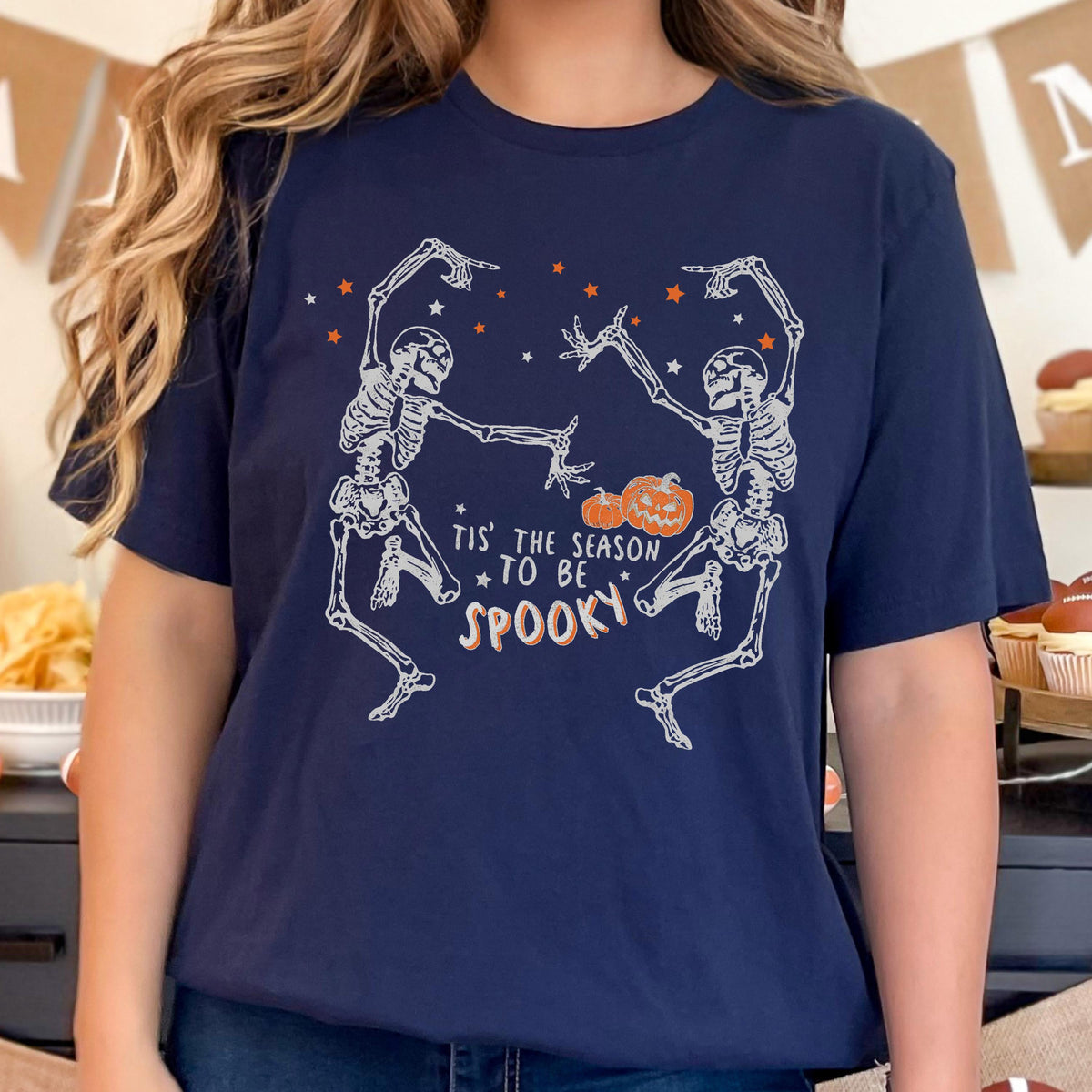 May include: Navy blue t-shirt featuring a Halloween design. Two dancing skeletons are depicted with orange stars and a pumpkin. The text reads "Tis' the season to be spooky" in white and orange.