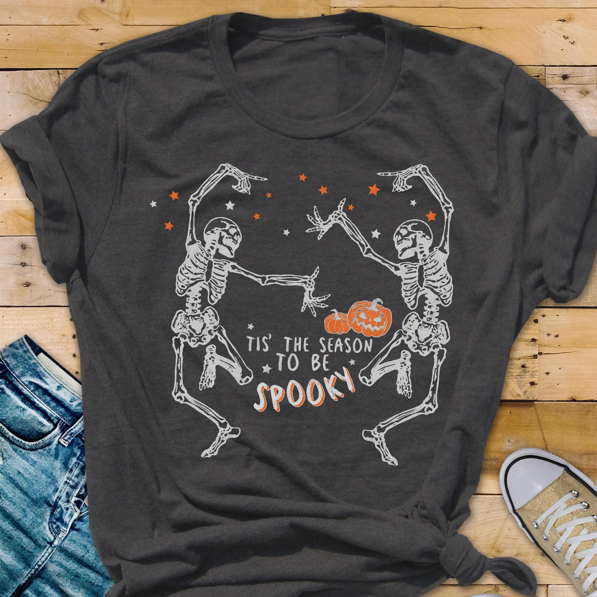 May include: Dark gray t-shirt featuring a Halloween design. Two skeletons are dancing, with orange stars and pumpkins. The text reads "Tis the season to be spooky."
