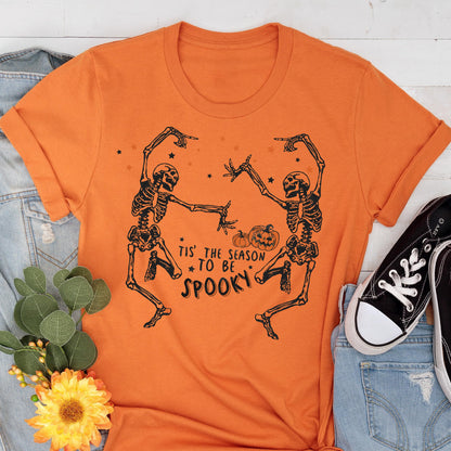 May include: Orange t-shirt featuring a black graphic of two dancing skeletons, pumpkins, and stars. The text "Tis' the season to be spooky" is printed below the skeletons. The shirt is displayed with denim and black sneakers.