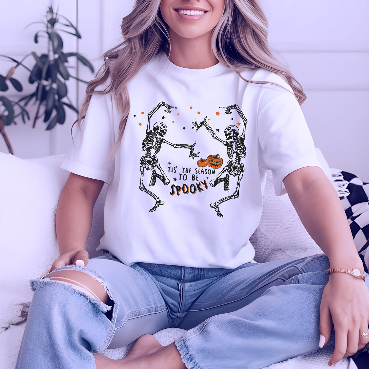 May include: White t-shirt featuring a Halloween design. The graphic includes two dancing skeletons, a pumpkin, and the text "Tis the season to be spooky" in black and orange. The shirt is a short-sleeved crew neck.