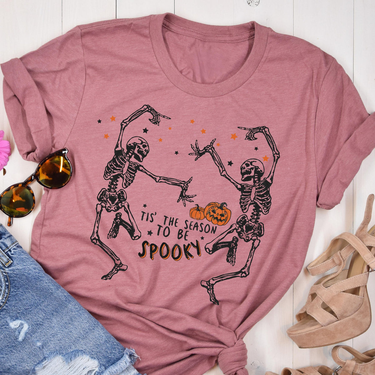 May include: Dusty rose-colored t-shirt featuring a black graphic of two dancing skeletons, pumpkins, and stars. The text "Tis' the season to be spooky" is printed below the skeletons. A casual, Halloween-themed tee.