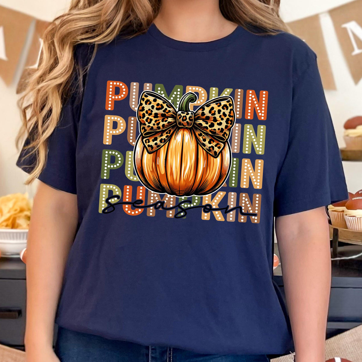 May include: Navy blue t-shirt featuring a large orange pumpkin with a leopard print bow. The words "PUMPKIN SEASON" are printed in a stacked design with orange, green, and white lettering. A festive fall-themed graphic.