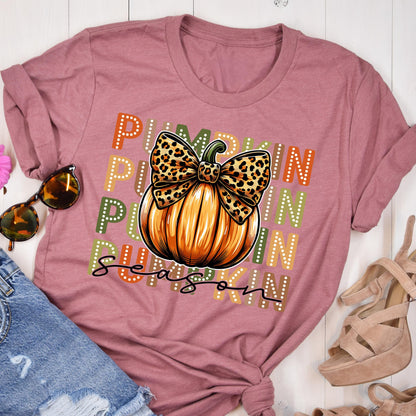 May include: A mauve-colored t-shirt featuring a pumpkin graphic with a leopard print bow. The words "Pumpkin Season" are printed on the shirt in a mix of orange, green, and white dotted letters. The shirt is styled with sunglasses and denim shorts.