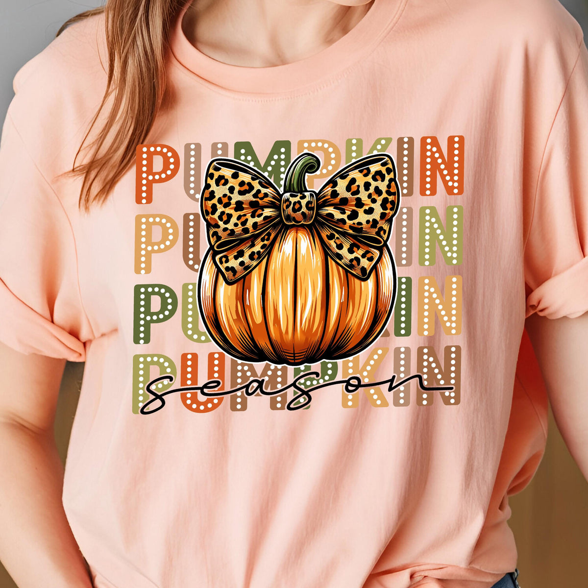 May include: Peach-colored t-shirt featuring a pumpkin graphic with a leopard print bow. The words "Pumpkin Season" are printed in a mix of colors. The design is centered on the front of the shirt.