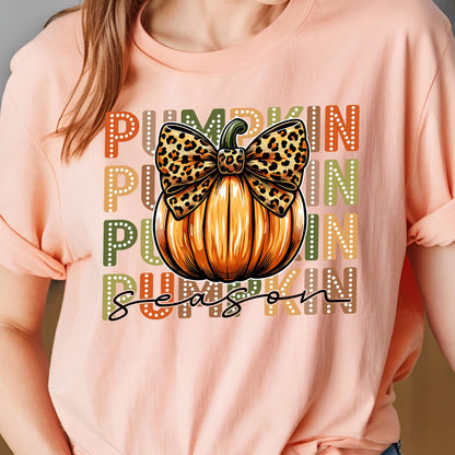 May include: Peach-colored t-shirt featuring a pumpkin graphic with a leopard print bow. The words "Pumpkin Season" are printed in a mix of colors. The design is centered on the front of the shirt.