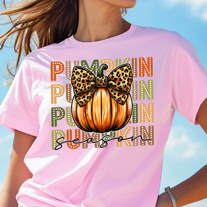 May include: Pink t-shirt featuring a large orange pumpkin with a leopard print bow. The word "PUMPKIN" is stacked vertically in orange, green, and yellow. The words "SEASON" are written in a script font.