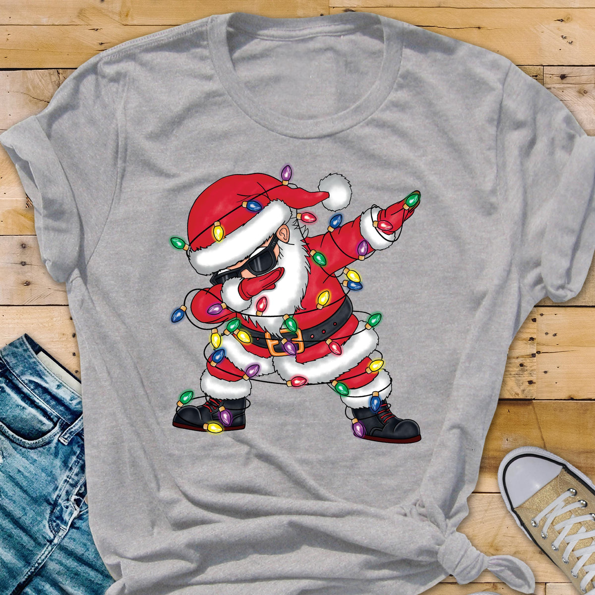 May include: A gray t-shirt featuring a graphic of Santa Claus dabbing, wearing sunglasses, a red suit, and a black belt. The Santa Claus graphic is decorated with colorful Christmas lights. The shirt is made of a soft material.