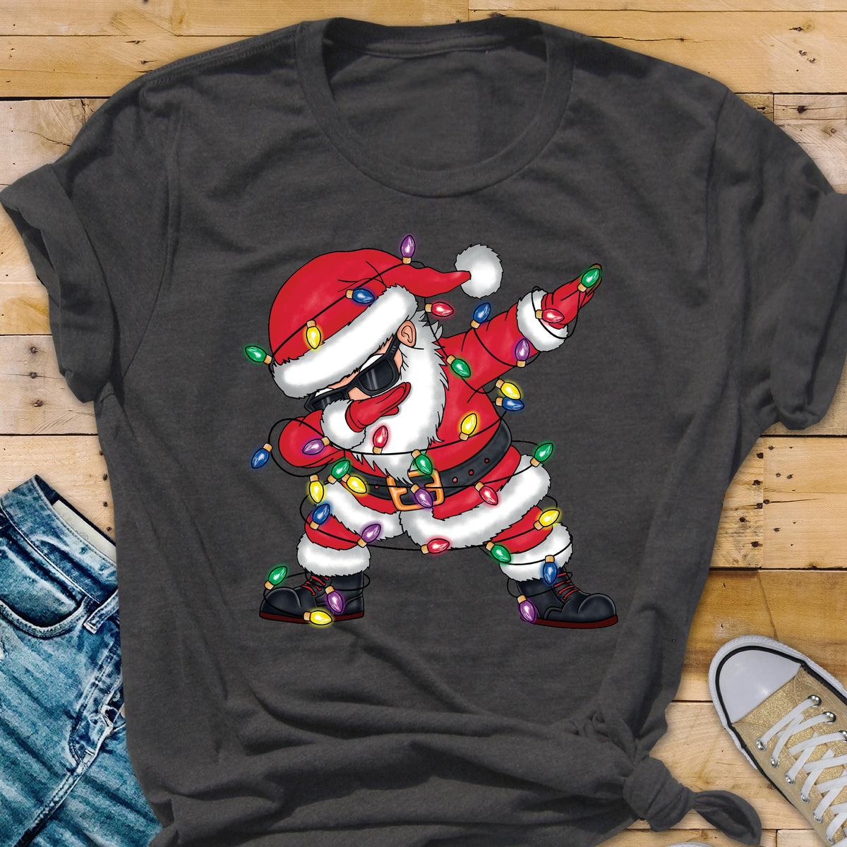 May include: Dark gray t-shirt featuring a cartoon Santa Claus dabbing, wearing sunglasses, and wrapped in Christmas lights. Santa is dressed in a red suit with a black belt and boots. The shirt is made of a soft material.