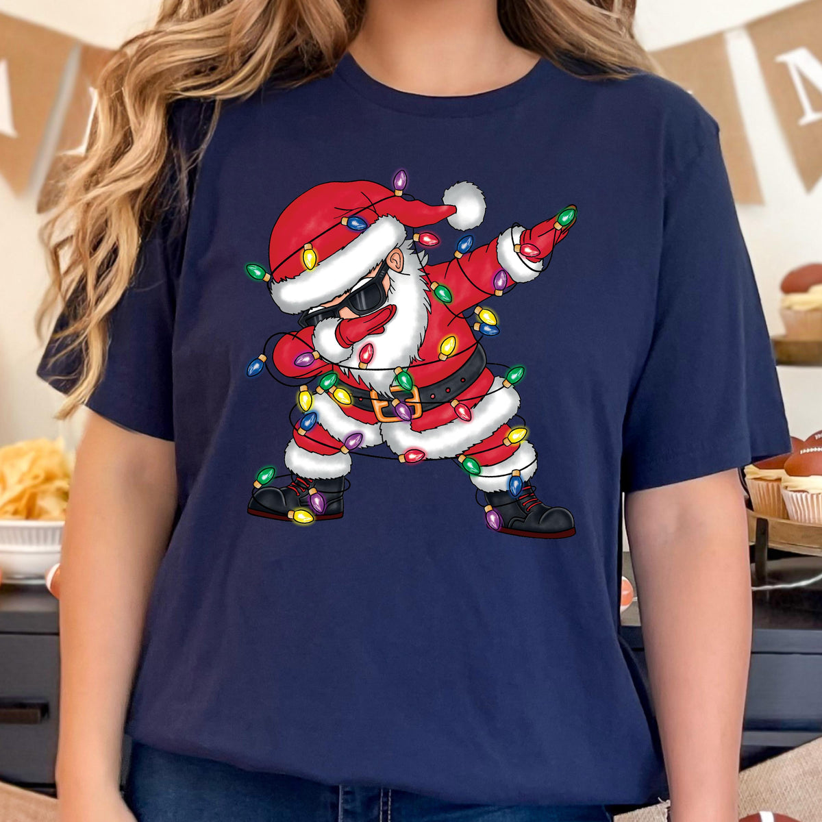 May include: Navy blue t-shirt featuring a cartoon Santa Claus dabbing. Santa is wearing sunglasses, a red suit, and a black belt. Colorful Christmas lights are wrapped around Santa's body. The shirt is a festive holiday design.