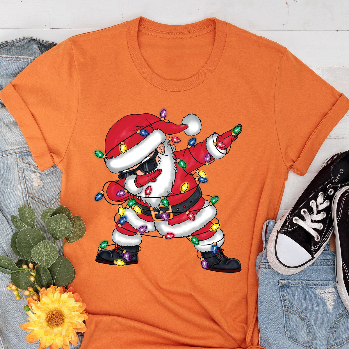 May include: Orange t-shirt featuring a graphic of Santa Claus dabbing, wearing sunglasses, and wrapped in Christmas lights. Santa is dressed in a red suit with a black belt and boots. The shirt is a crew neck style.