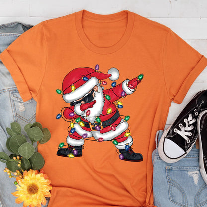 May include: Orange t-shirt featuring a graphic of Santa Claus dabbing, wearing sunglasses, and wrapped in Christmas lights. Santa is dressed in a red suit with a black belt and boots. The shirt is a crew neck style.