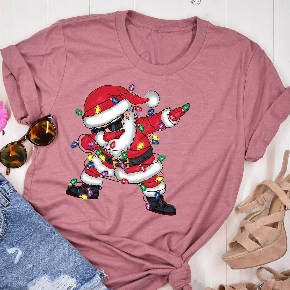 May include: A dusty rose t-shirt featuring a cartoon Santa Claus dabbing. Santa is wearing sunglasses, a red suit, and a hat, with a string of colorful Christmas lights wrapped around him. The design is centered on the shirt.