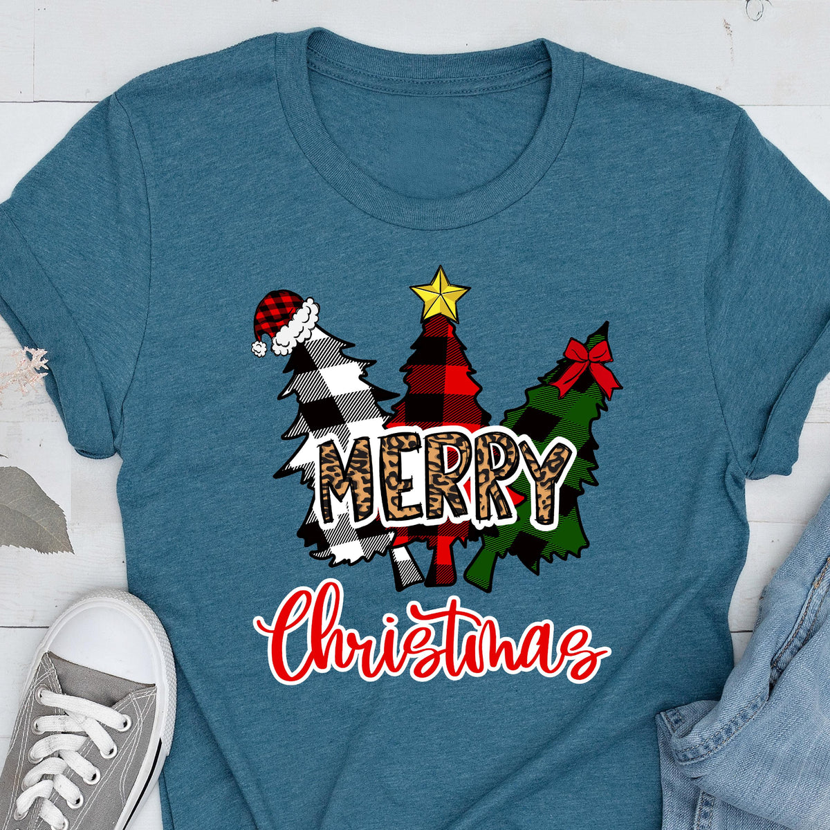 May include: A teal t-shirt featuring a graphic of three stylized Christmas trees in black and white buffalo plaid, red and black buffalo plaid, and green plaid. The word MERRY is in leopard print, and the word Christmas is in red script.