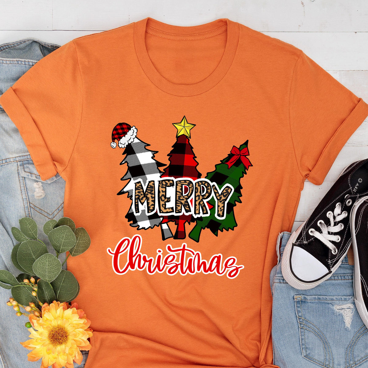 May include: An orange short-sleeved t-shirt featuring a graphic of three stylized Christmas trees. The trees have black and white plaid, red and black plaid, and green patterns. The word MERRY is in leopard print, and Christmas is in red script.