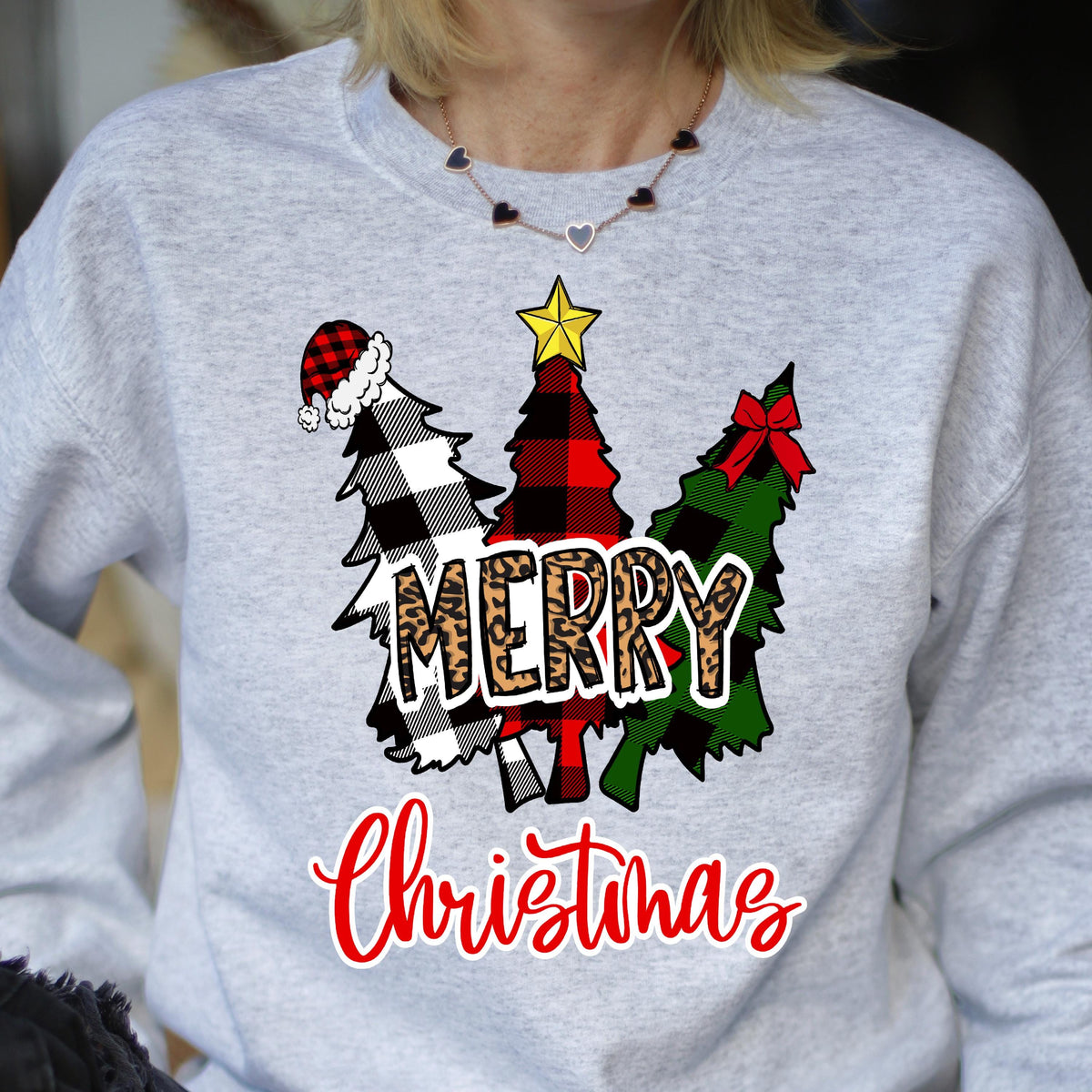 May include: A light gray sweatshirt featuring a graphic of three stylized Christmas trees. The trees have black and white buffalo plaid, red and black buffalo plaid, and green patterns. The word MERRY is in leopard print, and Christmas is in red.