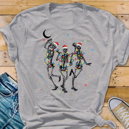 May include: A gray t-shirt featuring a festive design of three dancing skeletons wearing Santa hats and wrapped in colorful Christmas lights. A crescent moon and snowflakes are also included in the design.