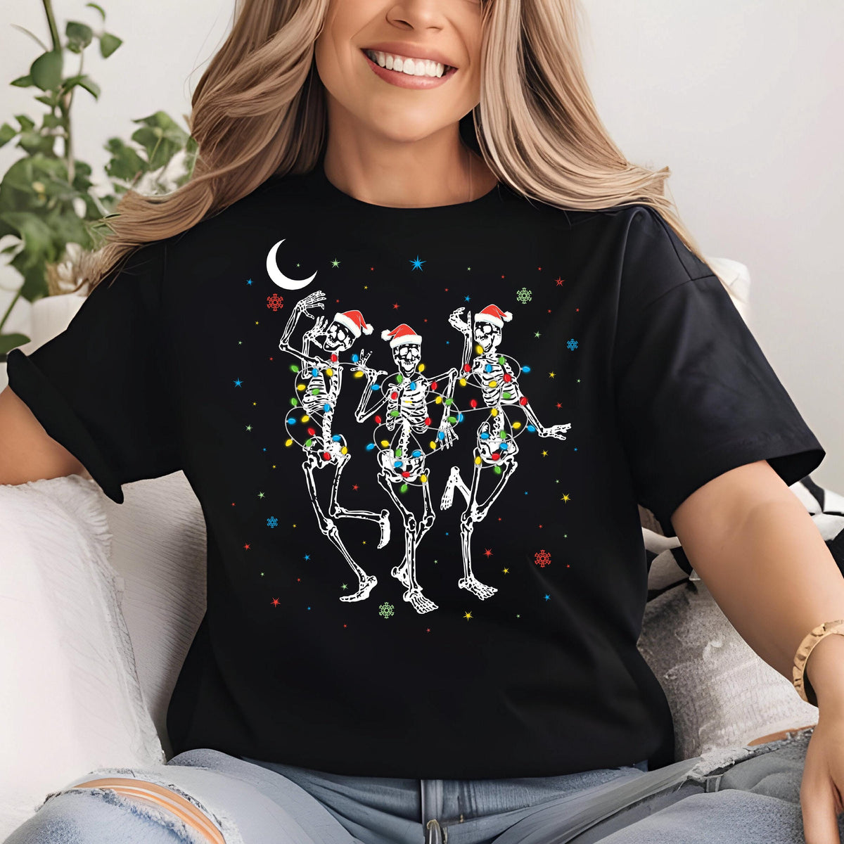 May include: Black t-shirt featuring a festive design of three dancing skeletons wearing Santa hats and wrapped in colorful Christmas lights. A crescent moon and snowflakes complete the holiday theme.
