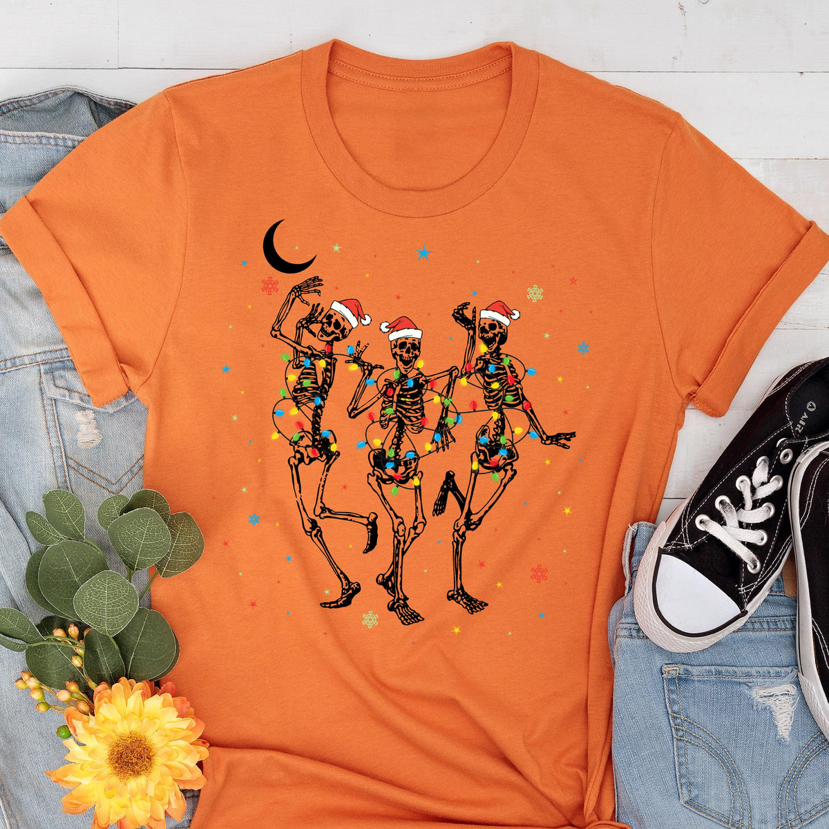 May include: Orange t-shirt featuring a graphic of three dancing skeletons adorned with Christmas lights and Santa hats. A crescent moon and colorful stars are also present in the design. The shirt is paired with blue jeans and black sneakers.
