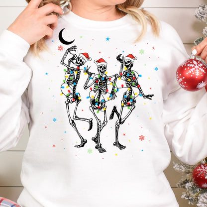May include: A white long-sleeve sweatshirt featuring a graphic of three dancing skeletons wearing red Santa hats. The skeletons are wrapped in colorful Christmas light strings. The design includes small red, blue, and green snowflakes and stars.