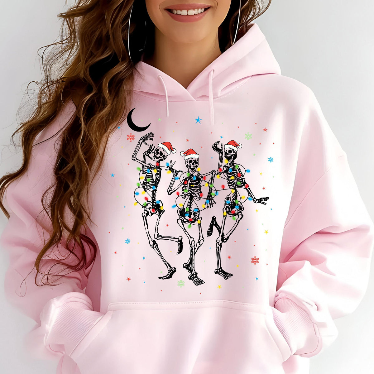 May include: A light pink hooded sweatshirt featuring a graphic print of three skeletons wearing red and white Santa hats. The skeletons are wrapped in colorful Christmas lights, surrounded by small stars, snowflakes, and a black crescent moon.