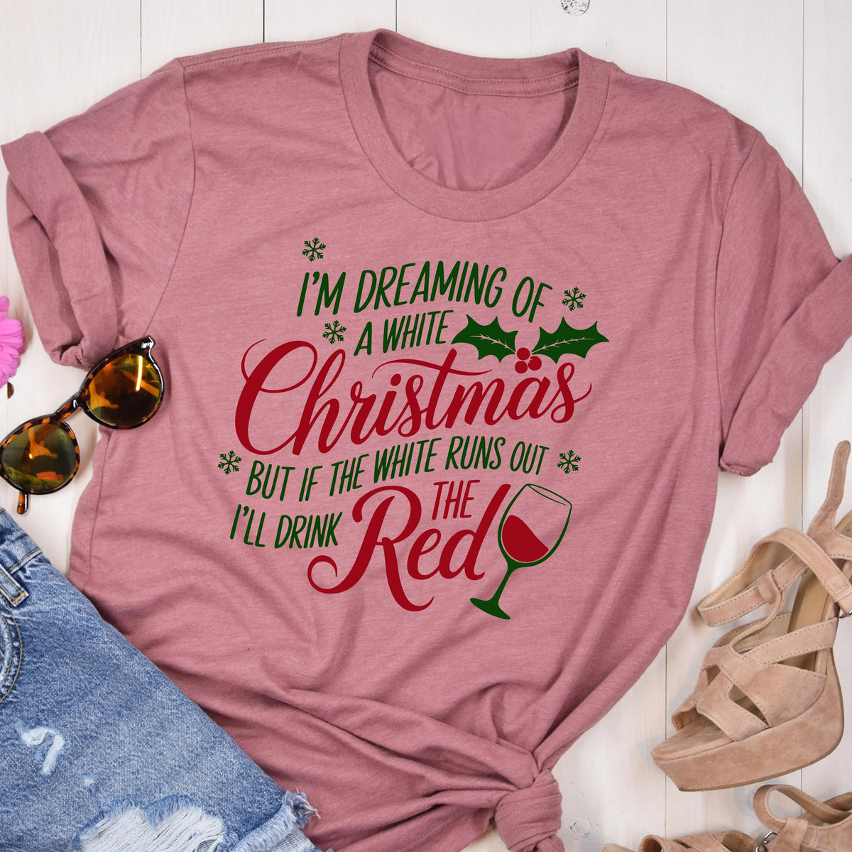 May include: A dusty rose-colored t-shirt with the text "I'M DREAMING OF A WHITE Christmas BUT IF THE WHITE RUNS OUT I'LL DRINK THE Red" in green and red. The design includes snowflakes, holly, and a wine glass.