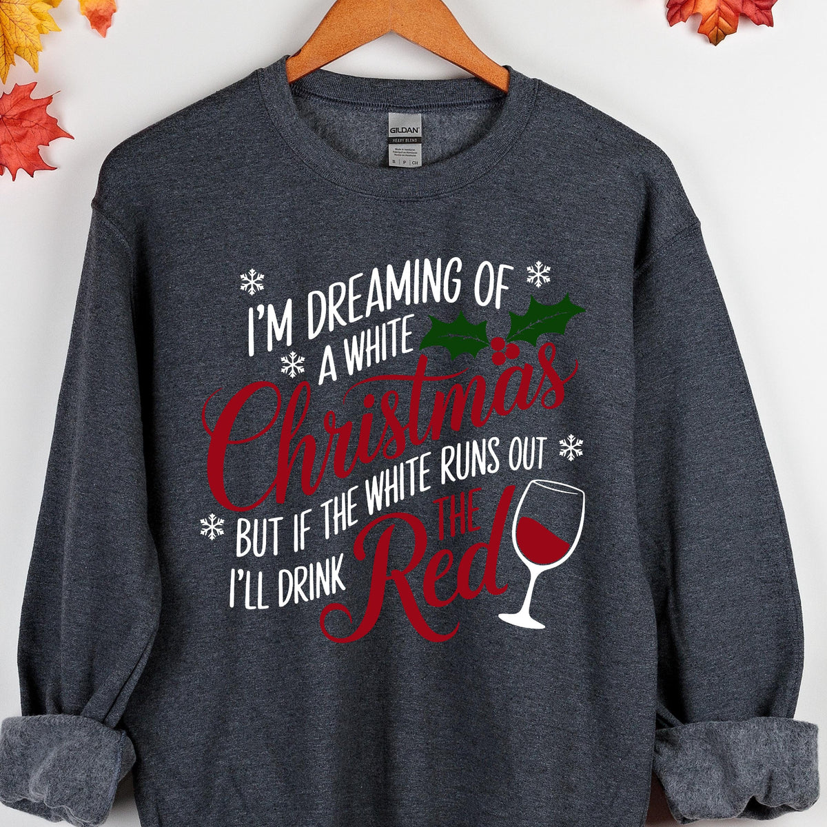 May include: A dark heather gray crewneck sweatshirt featuring white and red text that reads I'M DREAMING OF A WHITE CHRISTMAS BUT IF THE WHITE RUNS OUT I'LL DRINK THE RED. The design includes green holly leaves, red berries, snowflakes, and a wine glass.