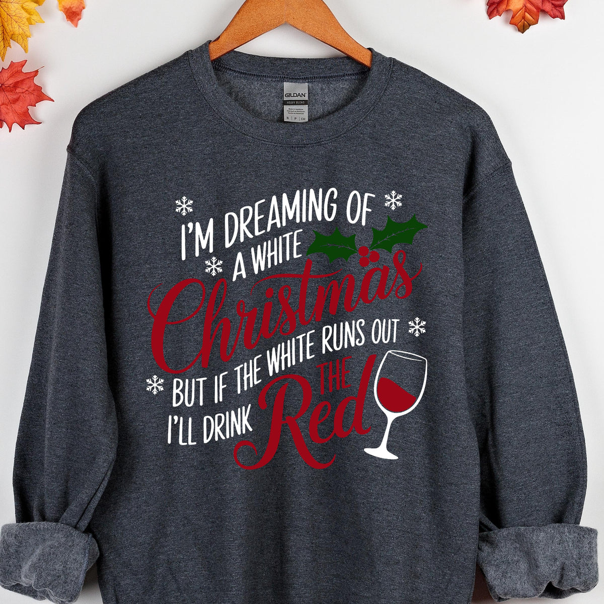 May include: A dark heather gray crewneck sweatshirt featuring white and red text that reads I'M DREAMING OF A WHITE CHRISTMAS BUT IF THE WHITE RUNS OUT I'LL DRINK THE RED. The design includes green holly leaves, red berries, snowflakes, and a wine glass.