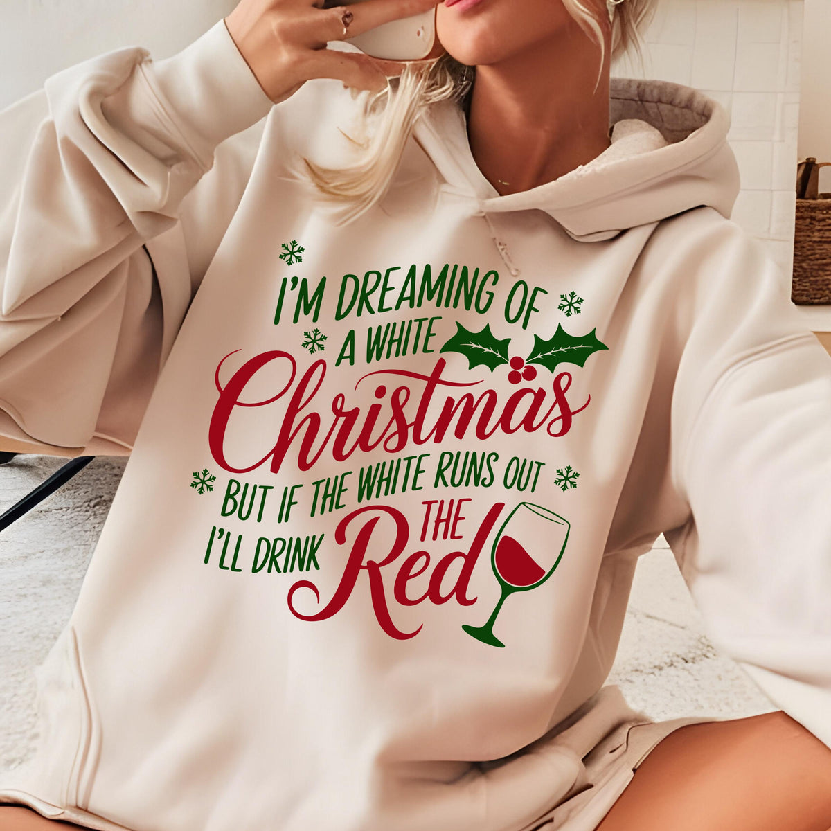 May include: A cream-colored hooded sweatshirt featuring a festive graphic with green and red text that reads: I'M DREAMING OF A WHITE CHRISTMAS BUT IF THE WHITE RUNS OUT I'LL DRINK THE RED. The design includes green holly leaves and a wine glass.