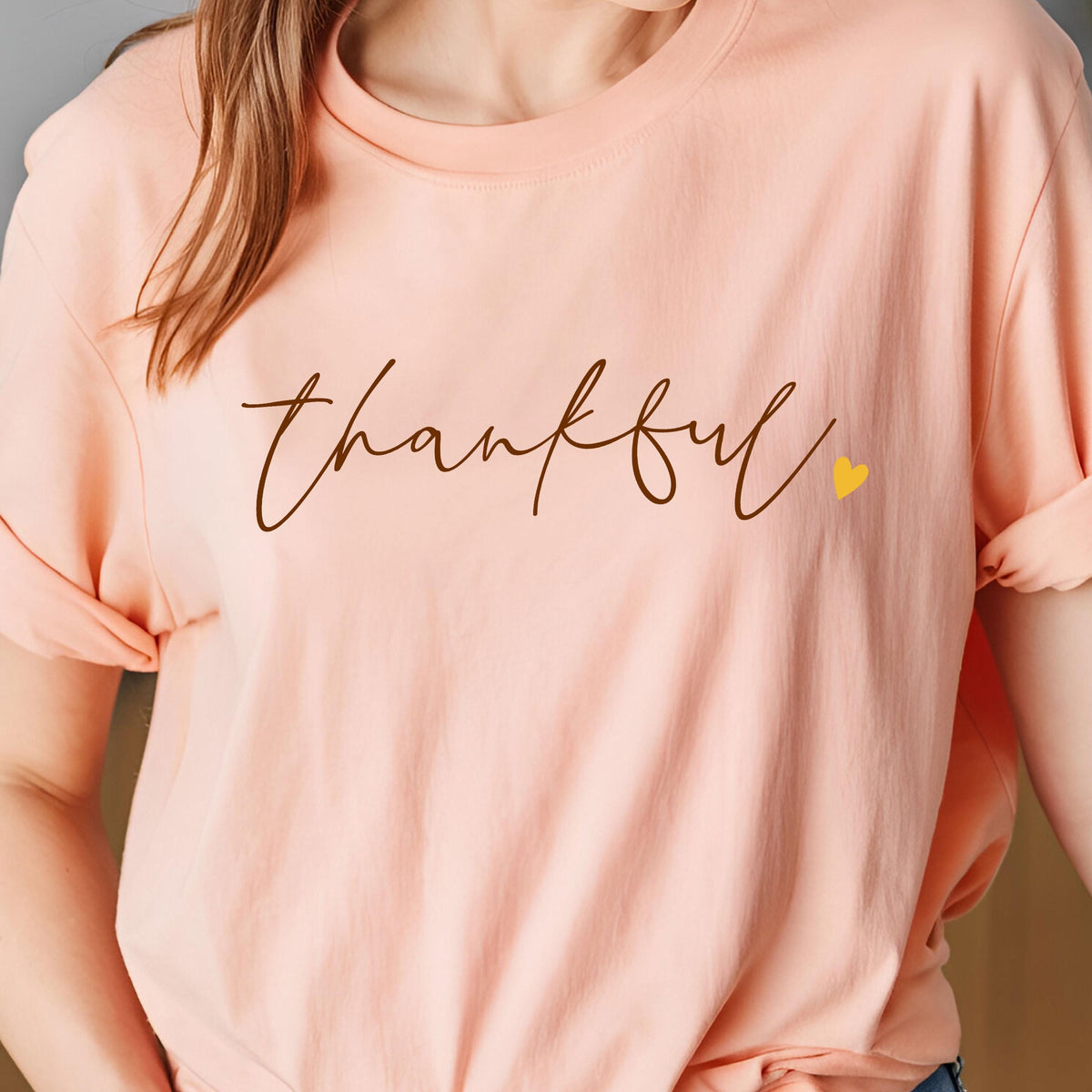 May include: A peach-colored t-shirt with the word "thankful" in a brown script font. A small yellow heart is next to the word. The sleeves are rolled up.