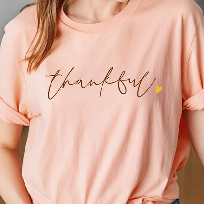 May include: A peach-colored t-shirt with the word "thankful" in a brown script font. A small yellow heart is next to the word. The sleeves are rolled up.