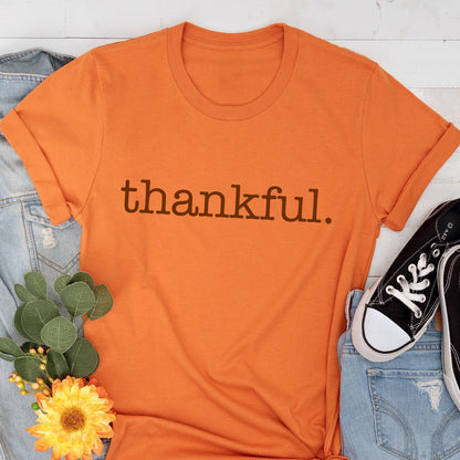 May include: Orange t-shirt with the word "thankful" printed in brown. The shirt is folded at the sleeves and is paired with blue jeans and black sneakers. A yellow flower and green leaves are placed near the shirt.