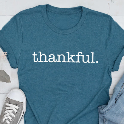 May include: Teal-colored t-shirt with the word "thankful" printed in white. The shirt is made of a soft material and has short sleeves. A pair of gray canvas sneakers and blue jeans are partially visible in the image.