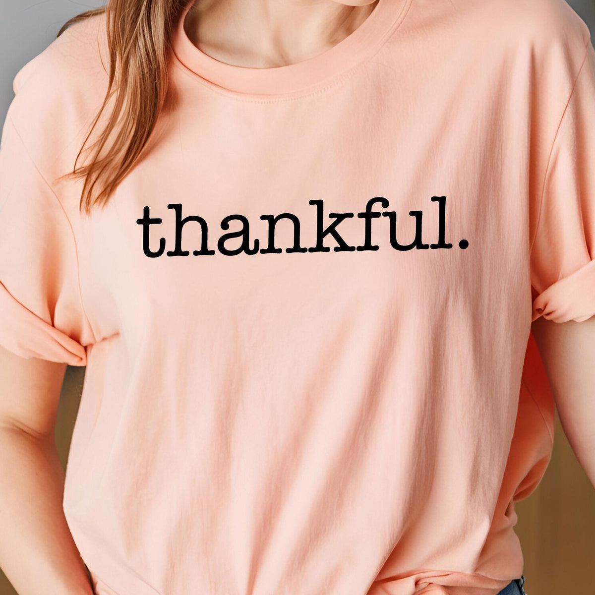 May include: A peach-colored t-shirt with the word "thankful." printed in black, centered on the front. The sleeves are rolled up. The shirt is made of a soft material.
