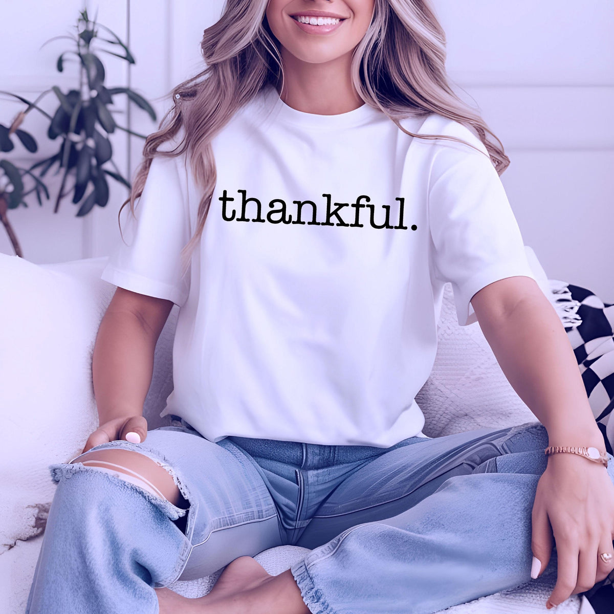 May include: White t-shirt with the word "thankful" printed in black. The shirt is short-sleeved and worn with light blue jeans. The person is sitting on a white couch.