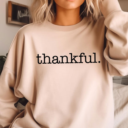 May include: A beige sweatshirt with the word "thankful" printed in black, centered on the front. The long-sleeved top is made of a soft material and is suitable for casual wear. The sweatshirt has a crew neck.