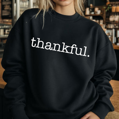 May include: Black sweatshirt with the word "thankful" printed in white, centered on the chest. The sweatshirt has a crew neck and long sleeves. The background is blurred, with a wooden surface in the foreground.