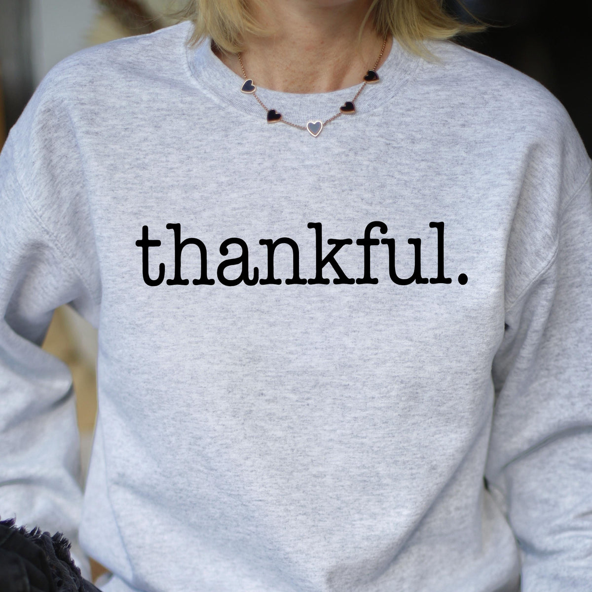 May include: A light gray sweatshirt with the word "thankful." printed in black. The sweatshirt is worn by a person, and a necklace with heart-shaped pendants is visible.