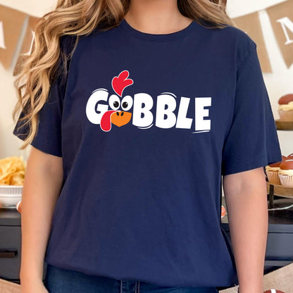 May include: A navy blue short-sleeve t-shirt featuring the white text GOBBLE printed across the chest. The letter O is replaced by a cartoon turkey face with a red wattle, a red comb, and large white eyes with black pupils, centered on the shirt.