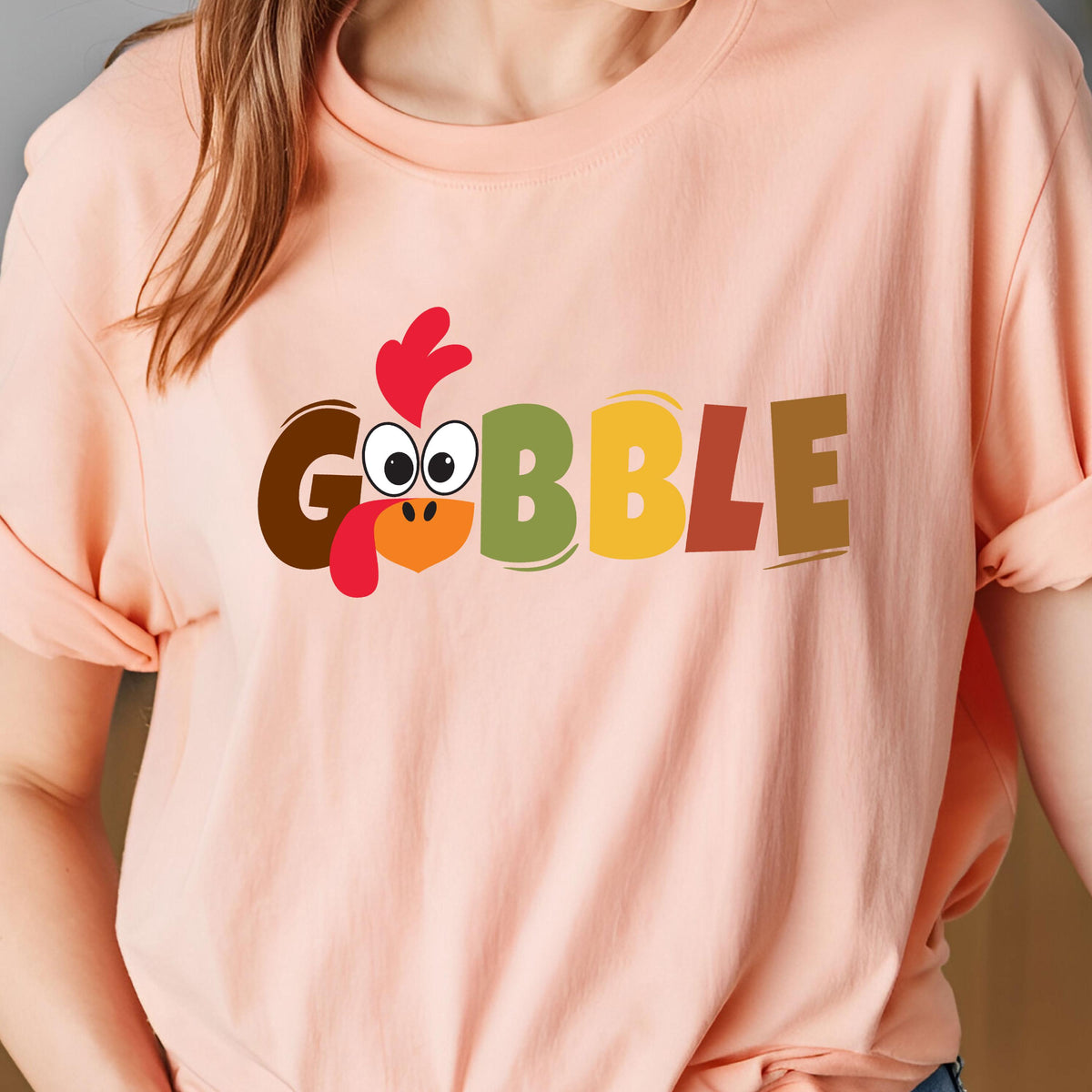 May include: A peach-colored t-shirt featuring the word GOBBLE printed in large, multi-colored block letters. The letter O is replaced by a cartoon turkey face with a red wattle, a bright orange beak, and large white eyes with black pupils.