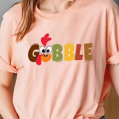 May include: A peach-colored t-shirt featuring the word GOBBLE printed in large, multi-colored block letters. The letter O is replaced by a cartoon turkey face with a red wattle, a bright orange beak, and large white eyes with black pupils.