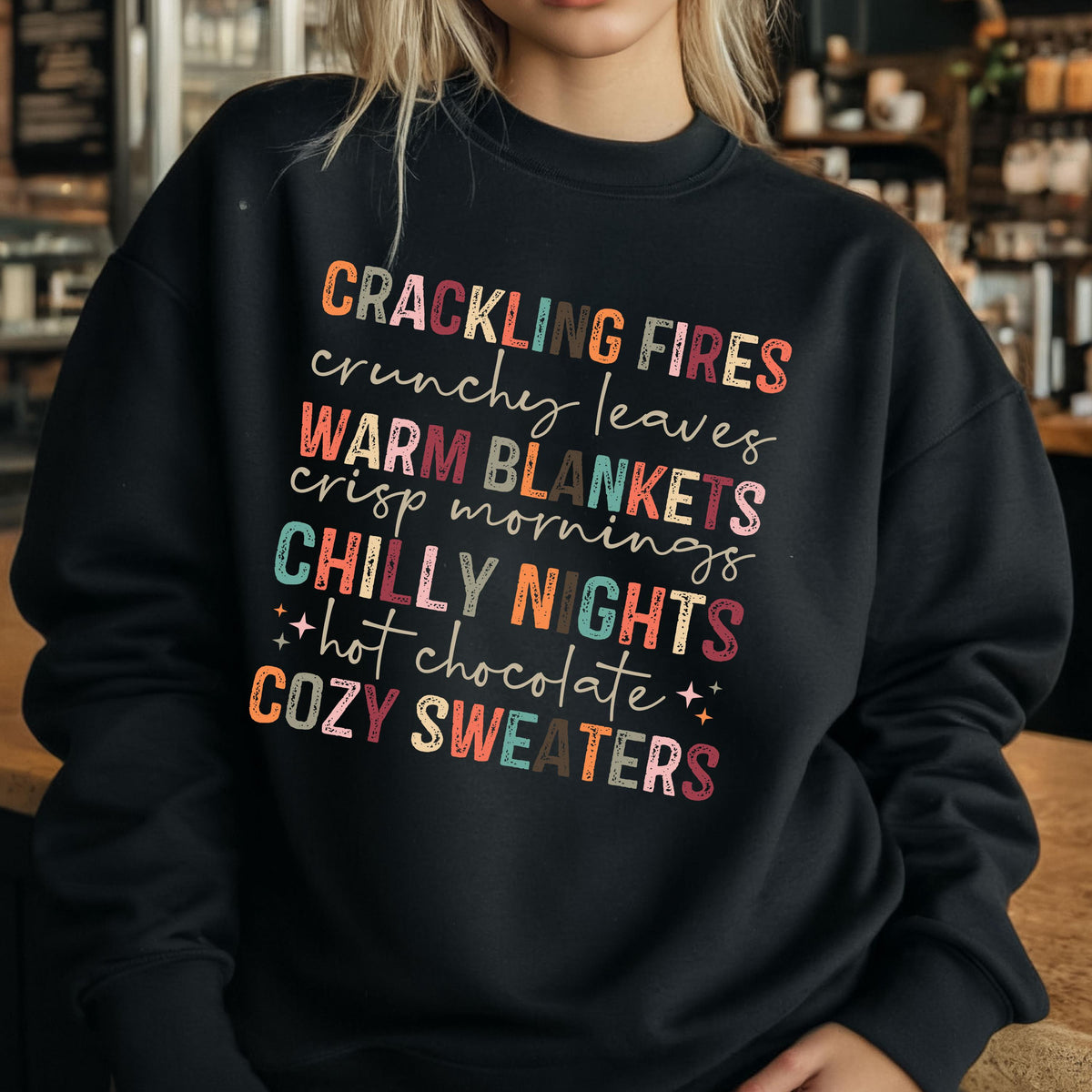 May include: Black sweatshirt with a colorful graphic print that reads "Crackling Fires, Crunchy Leaves, Warm Blankets, Crisp Mornings, Chilly Nights, Hot Chocolate, Cozy Sweaters".