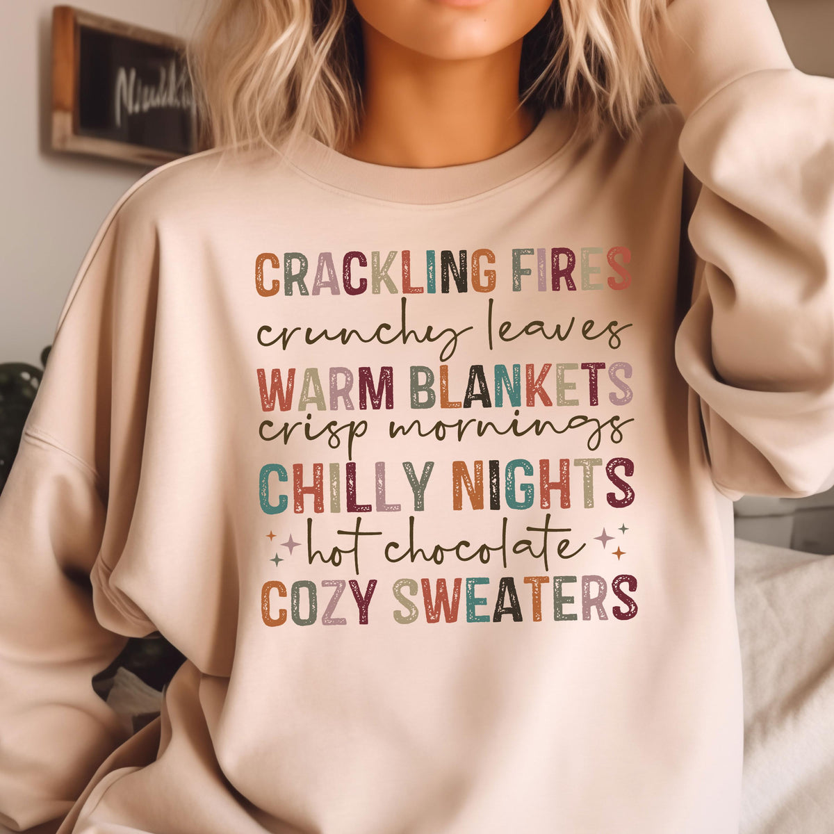May include: A beige sweatshirt with the text "CRACKLING FIRES crunchy leaves WARM BLANKETS crisp mornings CHILLY NIGHTS hot chocolate COZY SWEATERS" printed in colorful letters.