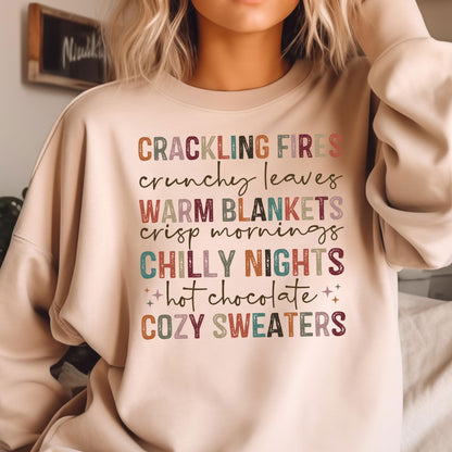 May include: A beige sweatshirt with the text "CRACKLING FIRES crunchy leaves WARM BLANKETS crisp mornings CHILLY NIGHTS hot chocolate COZY SWEATERS" printed in colorful letters.