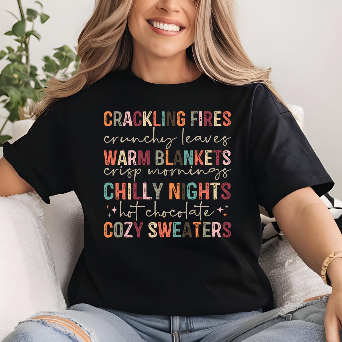 May include: Black t-shirt with colorful text: "CRACKLING FIRES, crunchy leaves, WARM BLANKETS, crisp mornings, CHILLY NIGHTS, hot chocolate, COZY SWEATERS." The text is in various fonts and colors, evoking a cozy autumn theme.