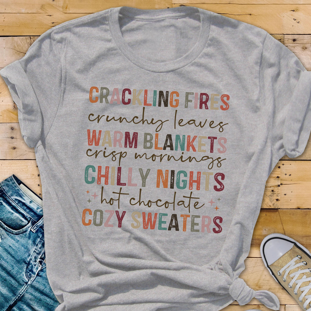 May include: A light gray t-shirt with the words "CRACKLING FIRES, CRUNCHY LEAVES, WARM BLANKETS, CRISP MORNINGS, CHILLY NIGHTS, hot chocolate, COZY SWEATERS" printed in various colors. The shirt is knotted at the bottom.