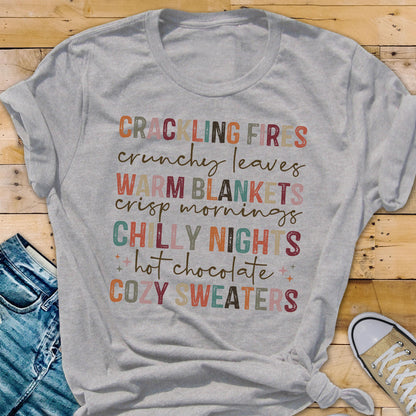 May include: A light gray t-shirt with the words "CRACKLING FIRES, CRUNCHY LEAVES, WARM BLANKETS, CRISP MORNINGS, CHILLY NIGHTS, hot chocolate, COZY SWEATERS" printed in various colors. The shirt is knotted at the bottom.